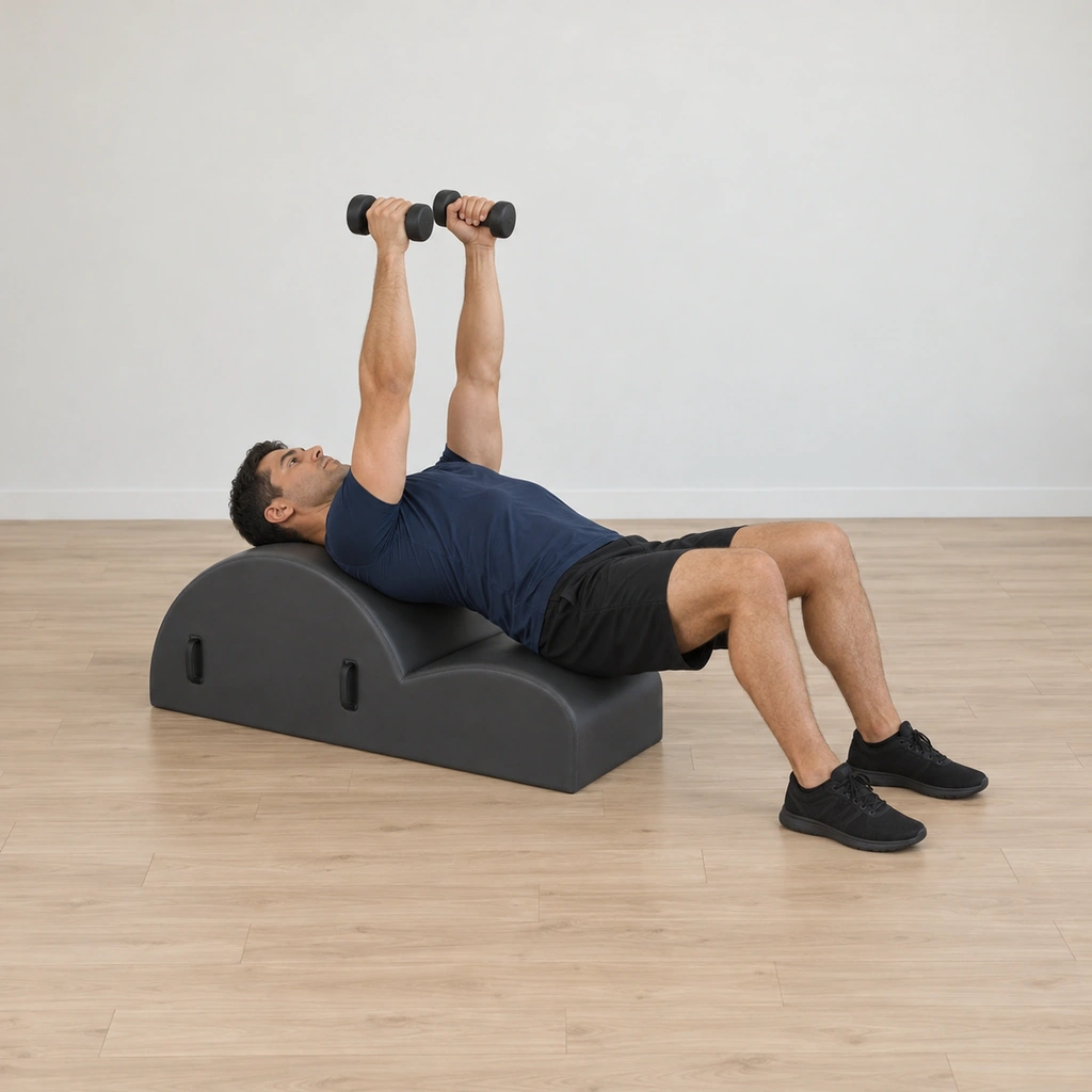 He leans back over the spine corrector's arc, one small weight in each hand held in front of his chest with the palms facing, his neck long and his upper back on the barrel.