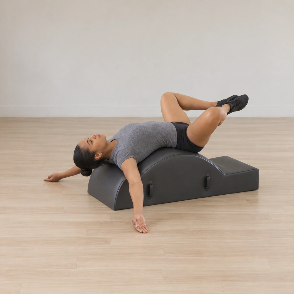 A woman lies draped back over the spine corrector arc, knees bent out wide in a frog with the heels hugged together and her gaze straight up.