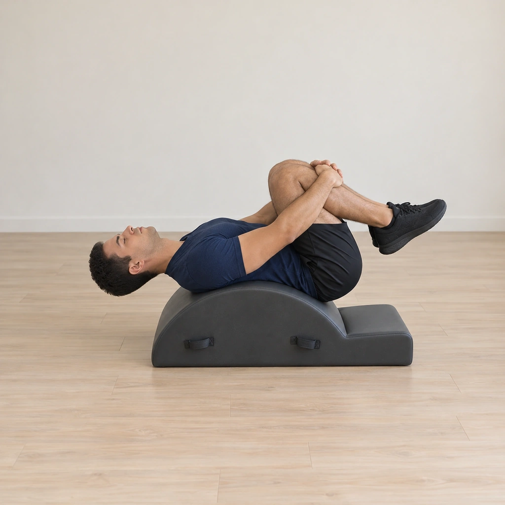 A man lies draped back over the spine corrector arc with his knees hugged together and folded, gazing up.