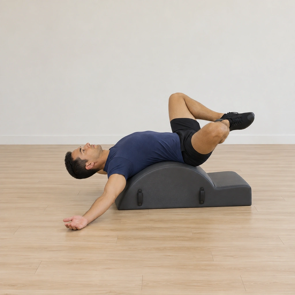 A man lies draped back over the spine corrector arc, knees bent wide in a frog with the heels hugged together and his gaze straight up.