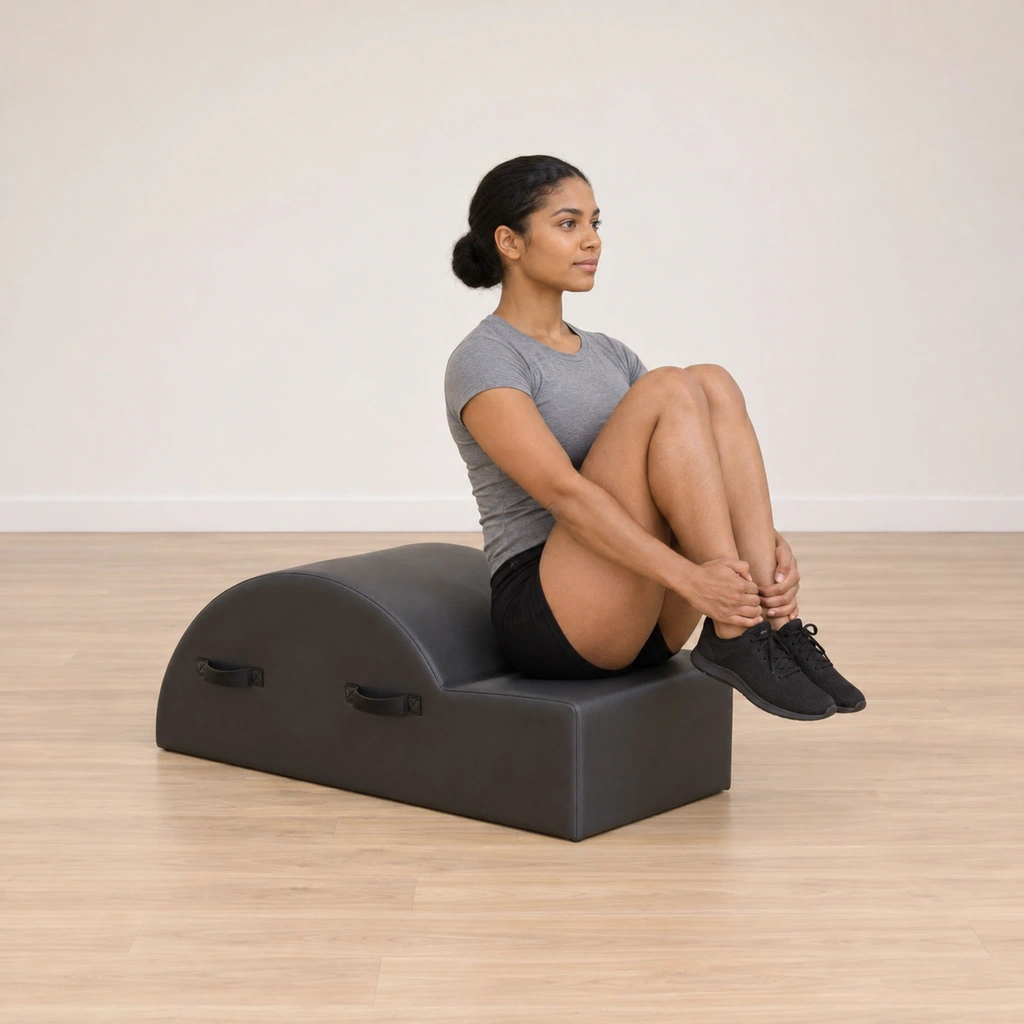 A woman sits in the spine corrector step crease with her back to the hump, knees drawn toward her chest, hands holding the fronts of her ankles, spine tall.