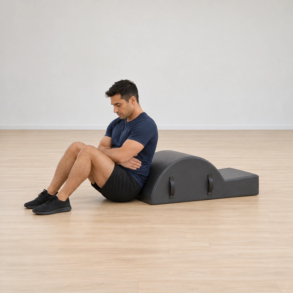 He sits tall on the floor just in front of the corrector's arc apex, knees bent, feet pressing the mat, his arms wrapped around his waist.
