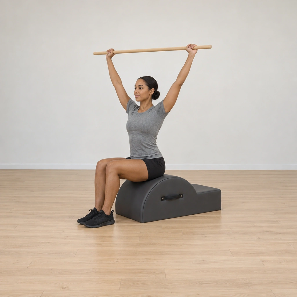 A woman sits tall on the corrector apex, feet planted a fist-distance apart pressing down, a long pole held overhead in wide straight arms slightly forward of her ears.