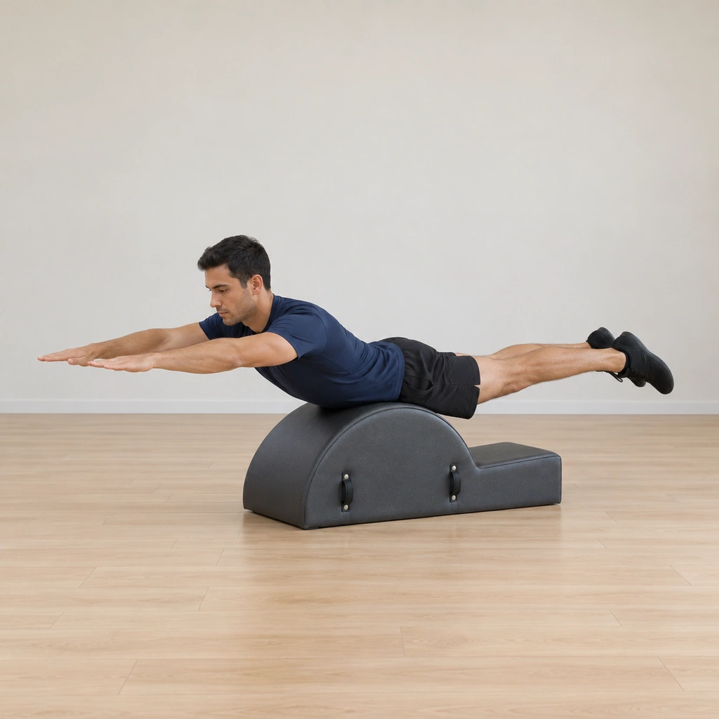 A man lies prone balanced on the apex of a spine corrector's arc, his chest and legs lifted into extension, thighs clear of the barrel, arms reached forward with the palms facing the floor.