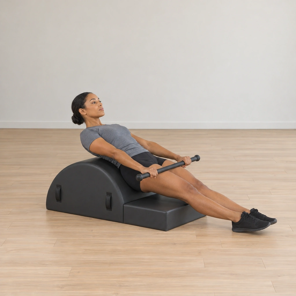 A woman reclines along the spine corrector arc facing away from it, her tailbone anchored at the wedge edge and legs long together, the weighted bar resting straight-armed along her thighs.