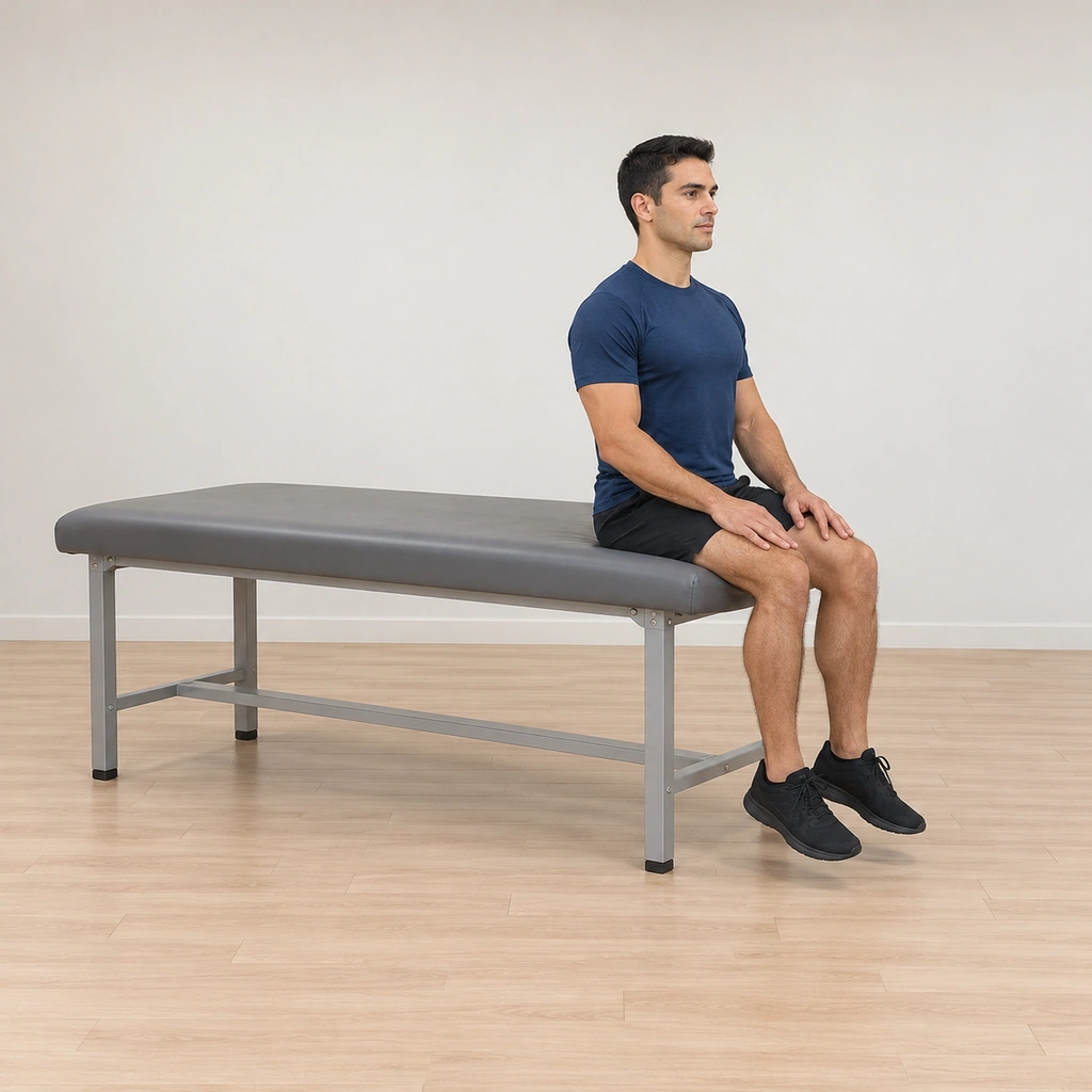He sits tall at the end of the exercise table with the mid-thigh line at the table edge, head lifted, hands resting, his trunk braced and his lower legs hanging down over the edge.