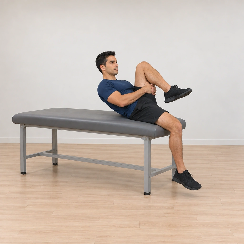He is leaning slightly back, his left knee drawn up off the table toward his chest with both hands folded under his left thigh, his trunk steady and his right leg still over the edge.