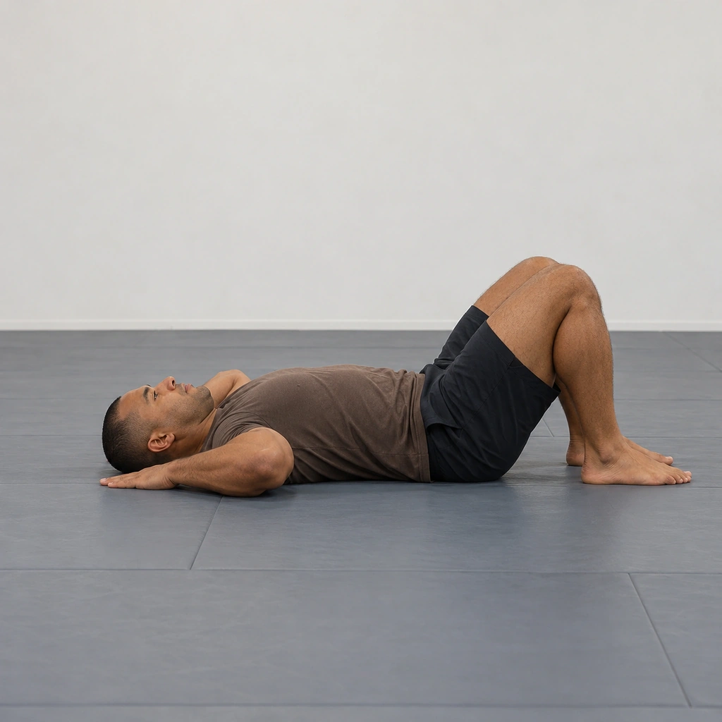 A man lying supine on a mat with knees bent, feet near his seat and hands flat on the mat beside his head, preparing to roll up into the bridge.